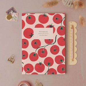 May include: A notebook with a tomato pattern. The cover features a repeating design of red tomatoes with green stems on a light pink background. The notebook has the text "NOTEBOOK" and "192 Plain Pages". A pink ruler and pencil are next to the notebook.