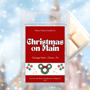 May include: A clear plastic clamshell package containing wax melts. The label reads "Christmas on Main" with "Orange Peel + Clove + Fir" below. The package is 5 oz. and hand-poured in Raleigh, NC.