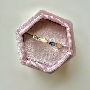 May include: A gold ring with a series of oval-shaped gemstones in white and blue, set in a gold band. The ring is displayed inside a pink velvet ring box. The gemstones are approximately 0.25 inches in length.