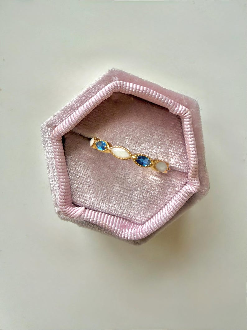 May include: A gold ring with a series of oval-shaped gemstones in white and blue, set in a gold band. The ring is displayed inside a pink velvet ring box. The gemstones are approximately 0.25 inches in length.