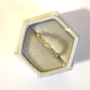 May include: A gold-toned ring with alternating oval and round gemstones in light blue and off-white. The ring is displayed inside a light gray velvet jewelry box.