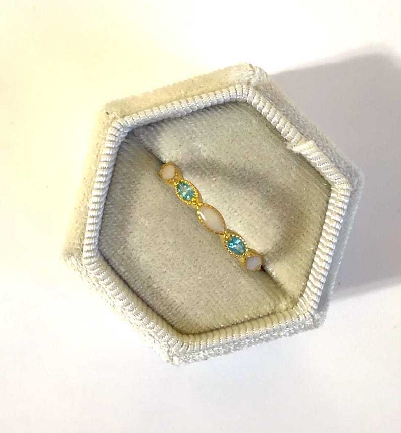 May include: A gold-toned ring with alternating oval and round gemstones in light blue and off-white. The ring is displayed inside a light gray velvet jewelry box.