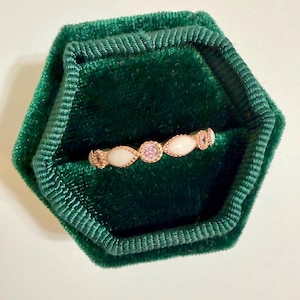 May include: A delicate gold ring with a series of oval-shaped white stones and a central pink gemstone. The ring is set in a dark green velvet jewelry box, creating a striking contrast. The ring's design is elegant and suitable for various occasions.
