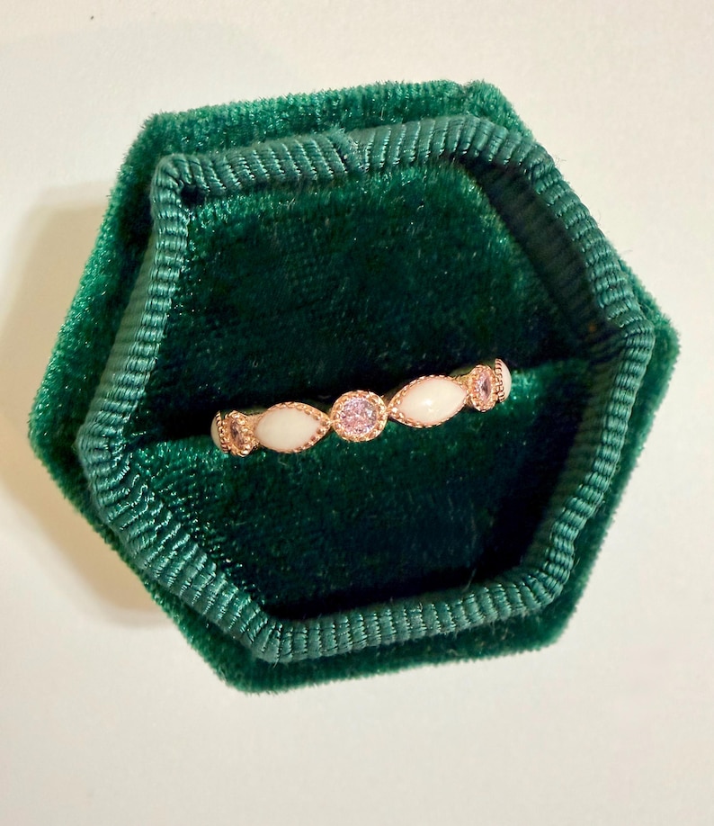 May include: A delicate gold ring with a series of oval-shaped white stones and a central pink gemstone. The ring is set in a dark green velvet jewelry box, creating a striking contrast. The ring's design is elegant and suitable for various occasions.