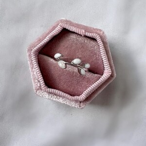 May include: A silver ring with a delicate branch design featuring white enamel leaves and a small, clear gemstone. The ring is displayed inside a pink velvet hexagonal jewelry box, set against a white background.