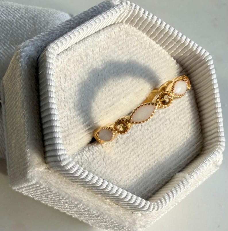 May include: A gold ring with a repeating pattern of oval and round gemstones. The ring is set in a white velvet box. The gemstones are a mix of white and light brown colors. The ring is a delicate piece of jewelry.
