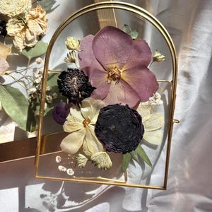 May include: A gold-framed glass display featuring pressed flowers. The arrangement includes a large purple flower, a dark black flower, and several smaller white and yellow blooms. The frame has an arched top and a rectangular base.