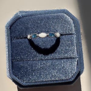 May include: A silver ring with a series of alternating blue and white gemstones, presented in a blue velvet ring box. The ring features a delicate design with oval and round stones, creating a subtle sparkle. The ring box is a deep blue color.