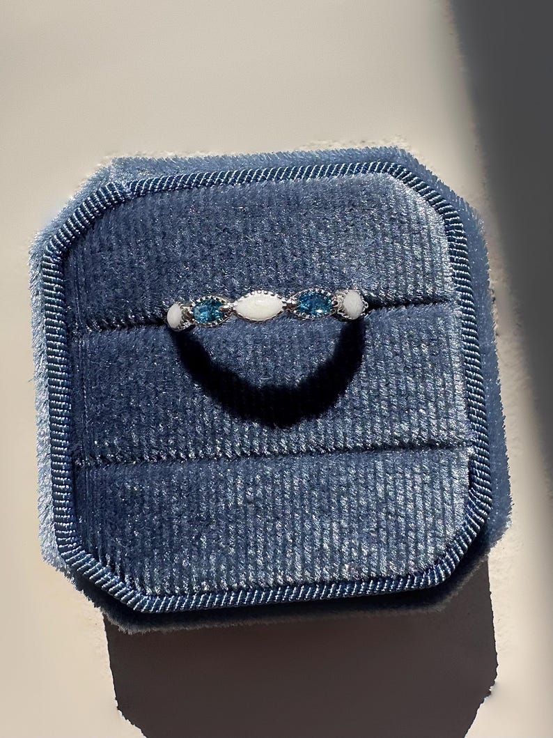 May include: A silver ring with a series of alternating blue and white gemstones, presented in a blue velvet ring box. The ring features a delicate design with oval and round stones, creating a subtle sparkle. The ring box is a deep blue color.