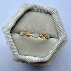 May include: A gold ring with a series of alternating pink and clear gemstones, set in a delicate band. The ring is displayed inside a gray velvet ring box, showcasing its intricate design and sparkle. The gemstones are oval-shaped.