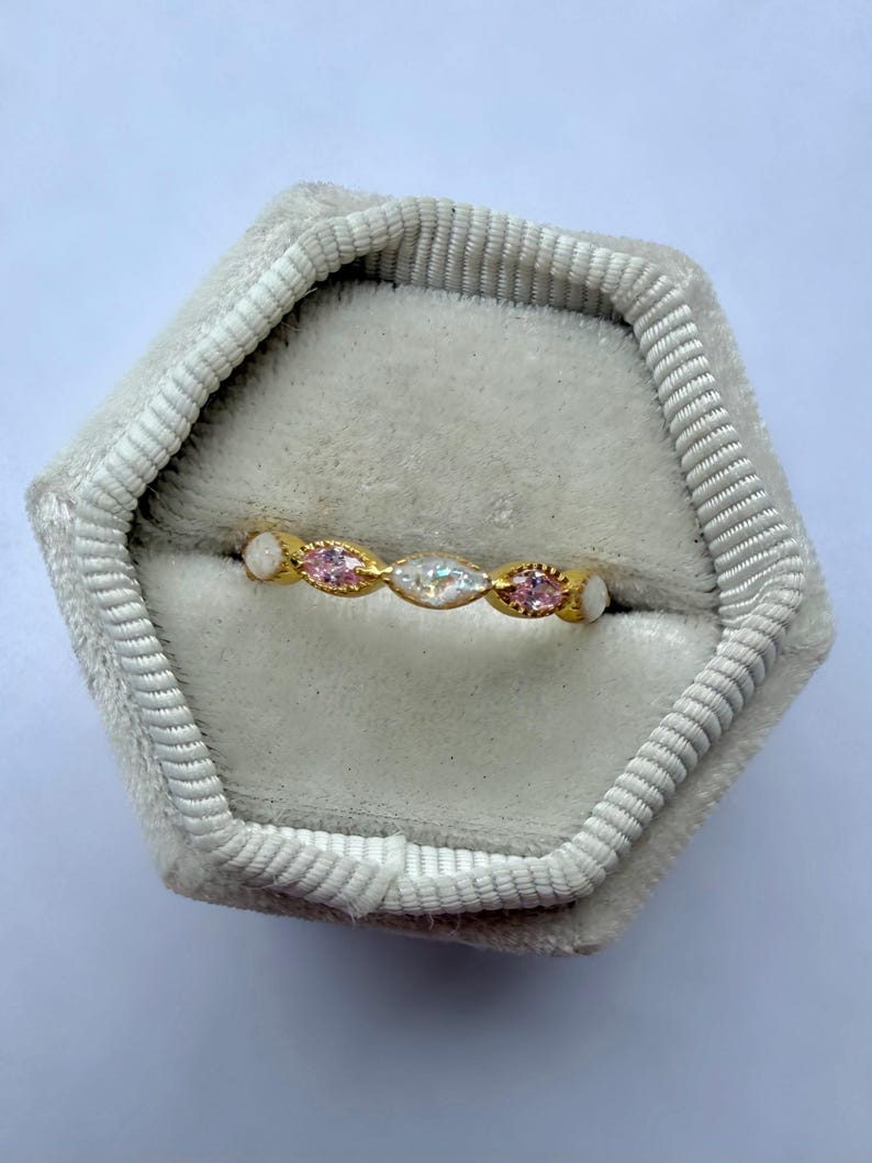 May include: A gold ring with a series of alternating pink and clear gemstones, set in a delicate band. The ring is displayed inside a gray velvet ring box, showcasing its intricate design and sparkle. The gemstones are oval-shaped.