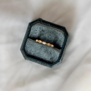 May include: A gold ring with small, white, oval-shaped stones and clear, round accent stones is displayed in a teal velvet ring box. The ring box is octagonal and sits on a white, textured surface.