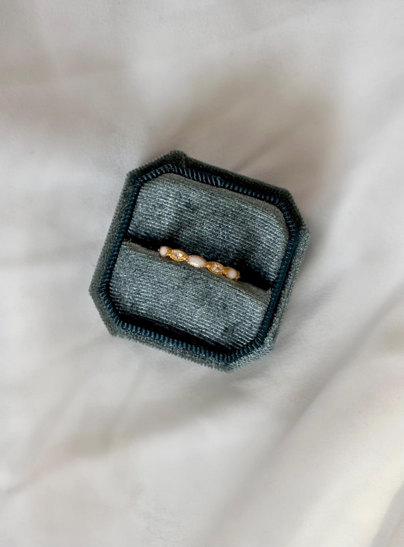 May include: A gold ring with small, white, oval-shaped stones and clear, round accent stones is displayed in a teal velvet ring box. The ring box is octagonal and sits on a white, textured surface.