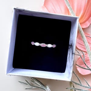 May include: A delicate ring with a silver band, featuring alternating pink and white gemstones. The ring is presented in a marble-patterned gift box with a black interior, set against a backdrop of pink flowers and greenery.