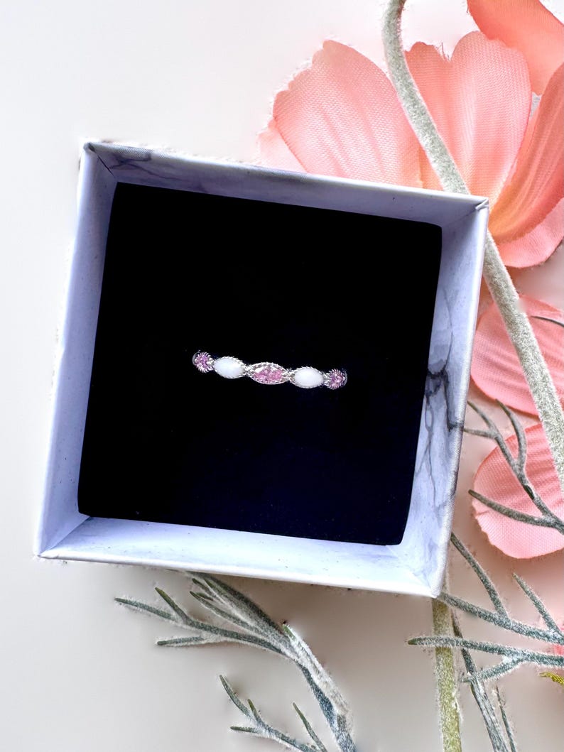 May include: A delicate ring with a silver band, featuring alternating pink and white gemstones. The ring is presented in a marble-patterned gift box with a black interior, set against a backdrop of pink flowers and greenery.