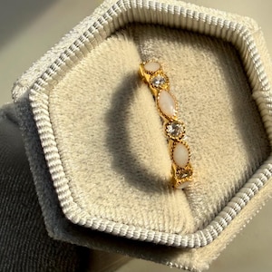 May include: A gold ring with alternating oval and round gemstones, displayed in a light gray velvet jewelry box. The ring features a series of white and clear stones. The jewelry box is hexagonal and has a soft, textured appearance.