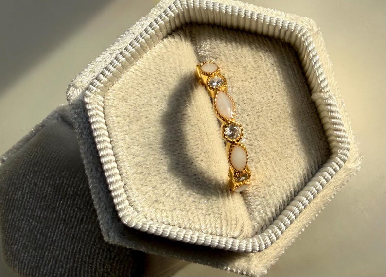 May include: A gold ring with alternating oval and round gemstones, displayed in a light gray velvet jewelry box. The ring features a series of white and clear stones. The jewelry box is hexagonal and has a soft, textured appearance.