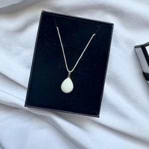 May include: A silver chain necklace featuring a smooth, white, teardrop-shaped pendant. The necklace is displayed inside a black rectangular jewelry box with a velvet-like interior lining, showcasing the contrast between the metal and stone.