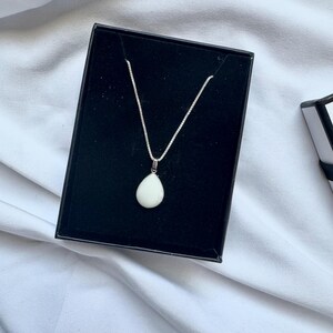 May include: A silver chain necklace featuring a smooth, white, teardrop-shaped pendant. The necklace is displayed inside a black rectangular jewelry box with a velvet-like interior lining, showcasing the contrast between the metal and stone.