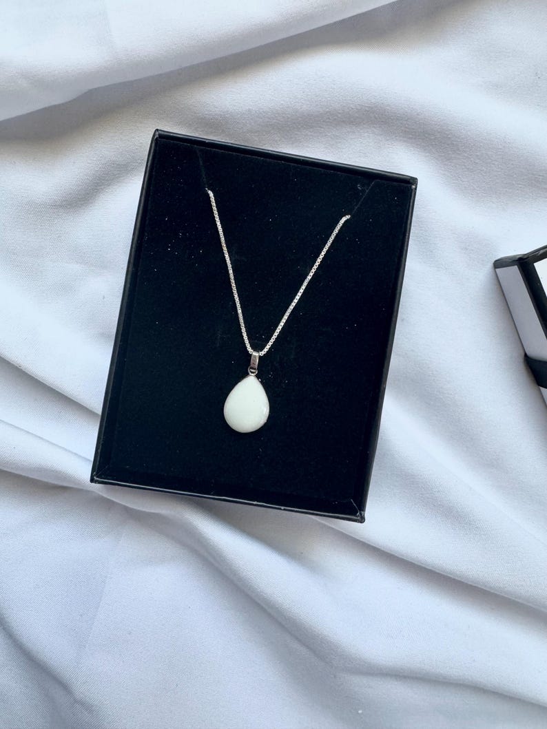 May include: A silver chain necklace featuring a smooth, white, teardrop-shaped pendant. The necklace is displayed inside a black rectangular jewelry box with a velvet-like interior lining, showcasing the contrast between the metal and stone.