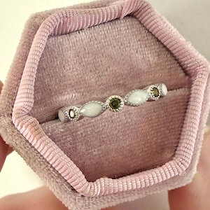 May include: A delicate silver ring with alternating white and green gemstones, presented in a pink velvet ring box. The ring features oval-shaped white stones and round green stones, creating a subtle yet elegant design.