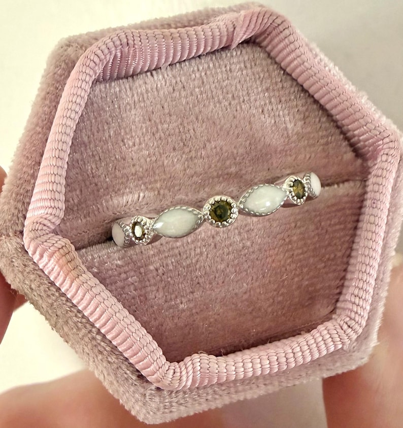 May include: A delicate silver ring with alternating white and green gemstones, presented in a pink velvet ring box. The ring features oval-shaped white stones and round green stones, creating a subtle yet elegant design.