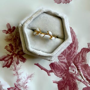 May include: A gold ring with a delicate branch design featuring white enamel accents, presented in a gray velvet ring box. The ring box is set against a backdrop of a floral patterned fabric.