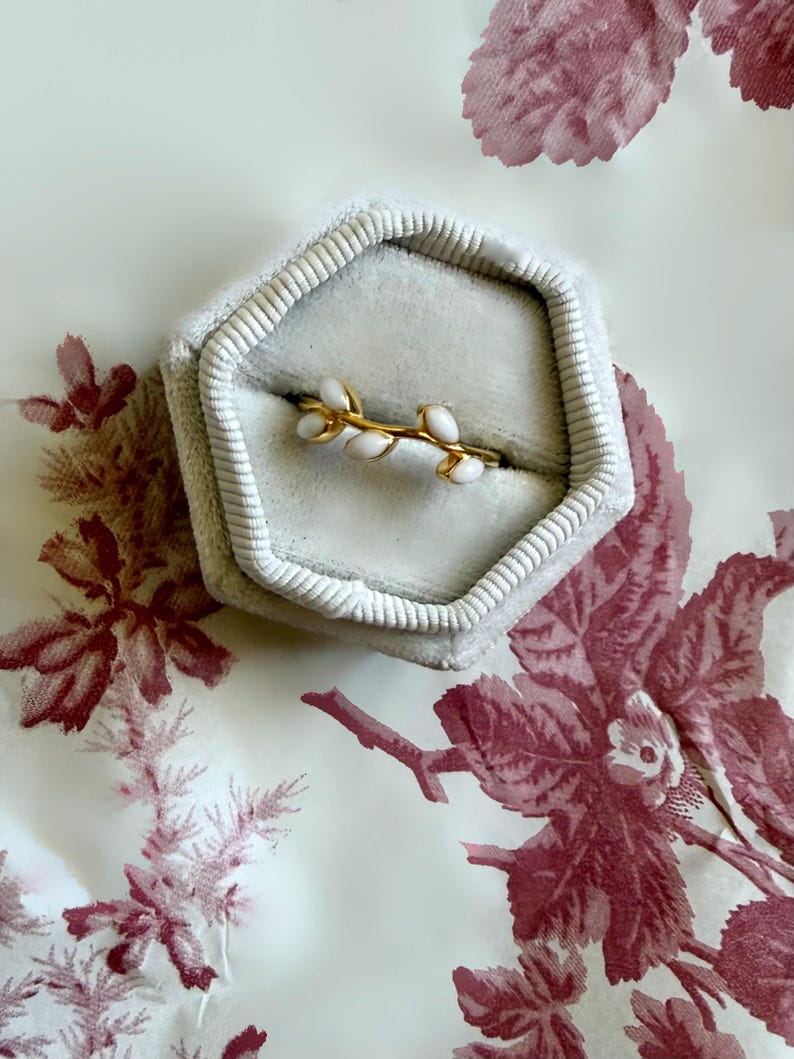 May include: A gold ring with a delicate branch design featuring white enamel accents, presented in a gray velvet ring box. The ring box is set against a backdrop of a floral patterned fabric.
