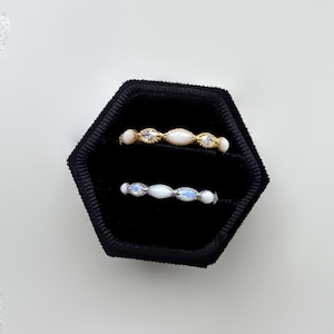 May include: Two delicate rings displayed in a black velvet box. One ring is gold with white and clear stones. The other ring is silver with white and blue stones. The rings are set against a white background.