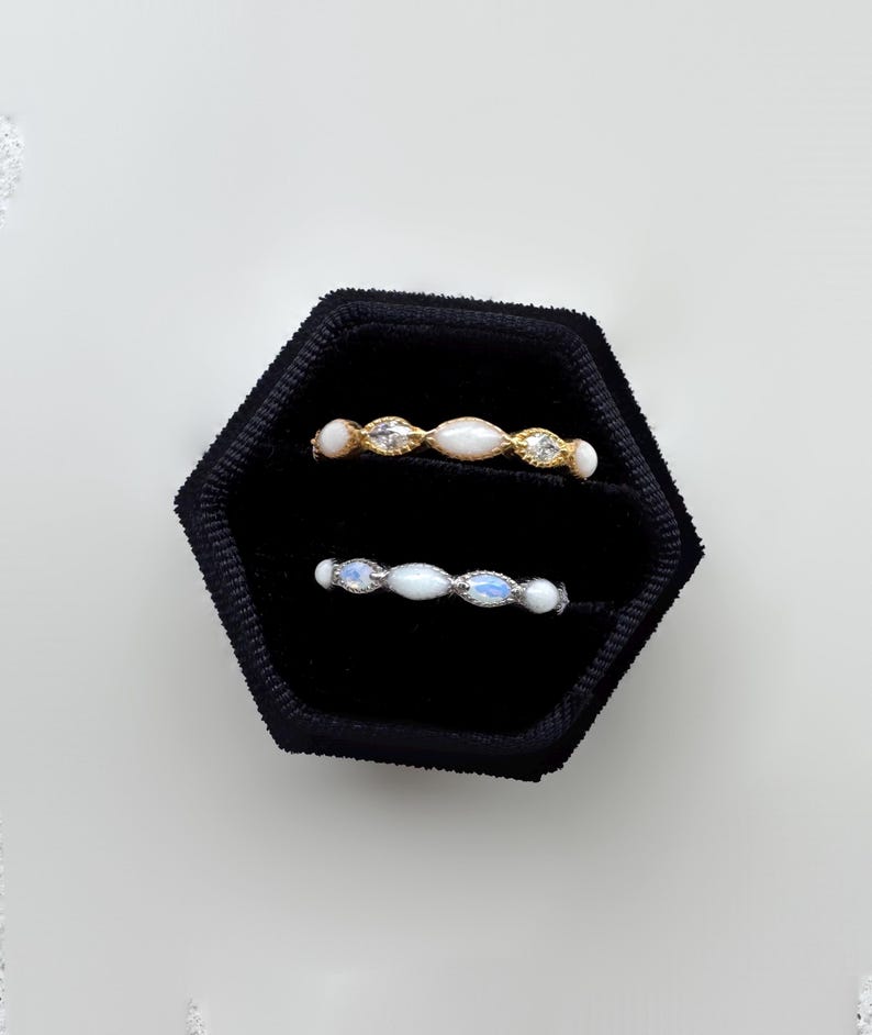 May include: Two delicate rings displayed in a black velvet box. One ring is gold with white and clear stones. The other ring is silver with white and blue stones. The rings are set against a white background.