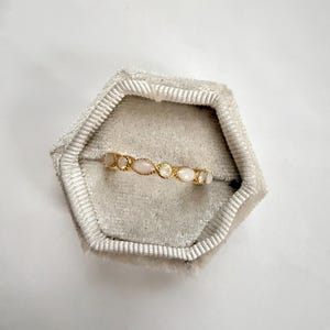 May include: A gold-colored ring with a series of oval and round, light-colored stones. The ring is set in a gold band and is displayed in a gray velvet ring box. The ring is a delicate piece of jewelry.