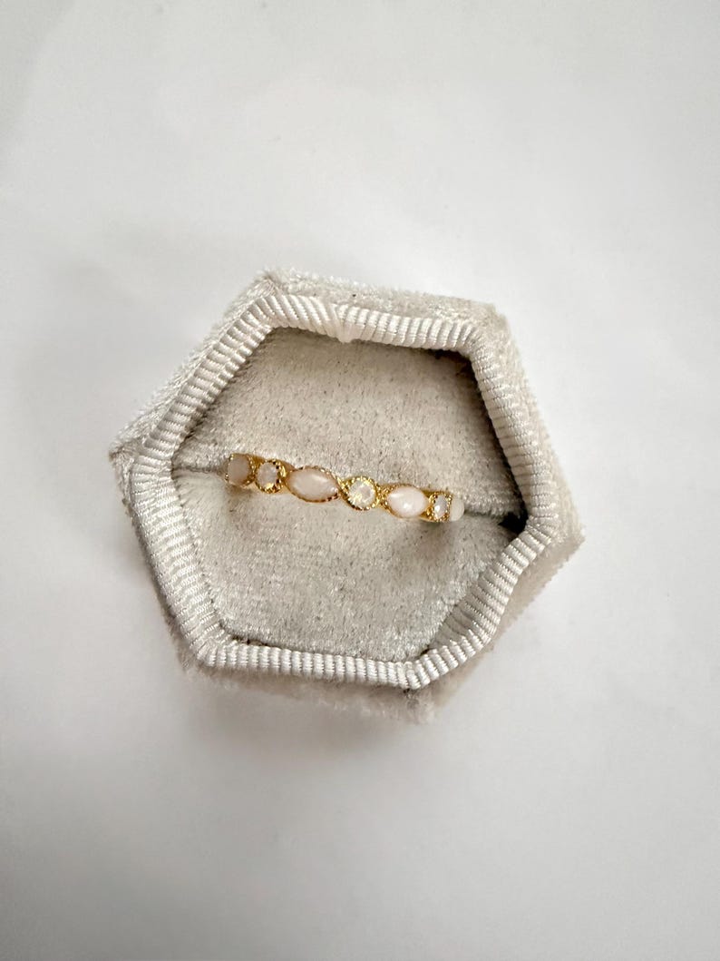 May include: A gold-colored ring with a series of oval and round, light-colored stones. The ring is set in a gold band and is displayed in a gray velvet ring box. The ring is a delicate piece of jewelry.