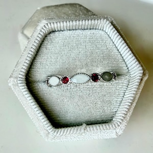 May include: A silver ring with a series of gemstones in a velvet ring box. The ring features a white, oval-shaped stone, two red stones, and a gray stone. The ring is set in a silver band and is displayed in a gray velvet ring box.