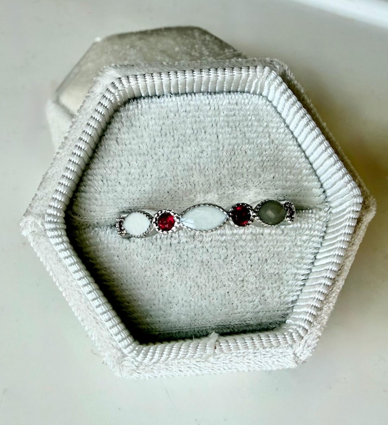 May include: A silver ring with a series of gemstones in a velvet ring box. The ring features a white, oval-shaped stone, two red stones, and a gray stone. The ring is set in a silver band and is displayed in a gray velvet ring box.