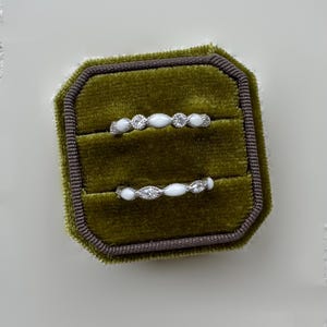 May include: Two delicate rings displayed in a green velvet ring box. Each ring features a series of small, clear gemstones and white, oval-shaped stones. The ring box is olive green with a dark brown trim.