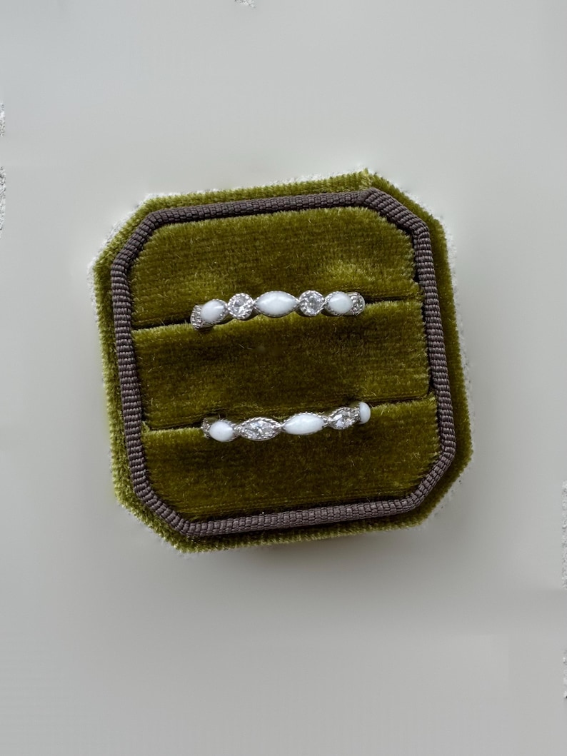 May include: Two delicate rings displayed in a green velvet ring box. Each ring features a series of small, clear gemstones and white, oval-shaped stones. The ring box is olive green with a dark brown trim.