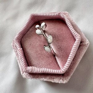 May include: A silver ring with a branch design, featuring white oval and round accents. The ring is displayed in a pink velvet hexagonal ring box, set against a white background.