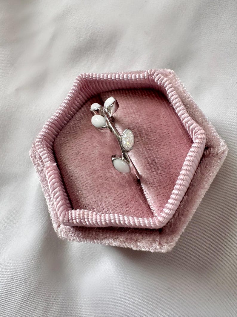May include: A silver ring with a branch design, featuring white oval and round accents. The ring is displayed in a pink velvet hexagonal ring box, set against a white background.