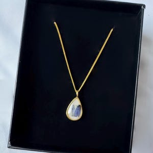 May include: A gold-toned necklace featuring a teardrop-shaped pendant with a white background and a small blue floral element encased inside. The pendant hangs from a delicate gold-toned chain, presented within a black rectangular jewelry box.