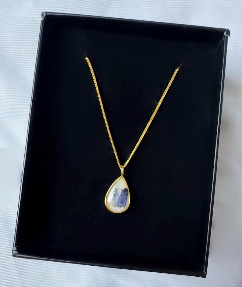May include: A gold-toned necklace featuring a teardrop-shaped pendant with a white background and a small blue floral element encased inside. The pendant hangs from a delicate gold-toned chain, presented within a black rectangular jewelry box.