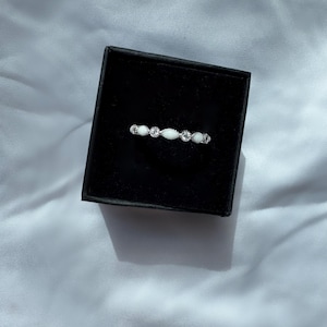 May include: A delicate silver ring with a series of small, clear gemstones and oval-shaped white stones. The ring is displayed inside a black jewelry box, set against a white fabric background. The ring's design is symmetrical and elegant.