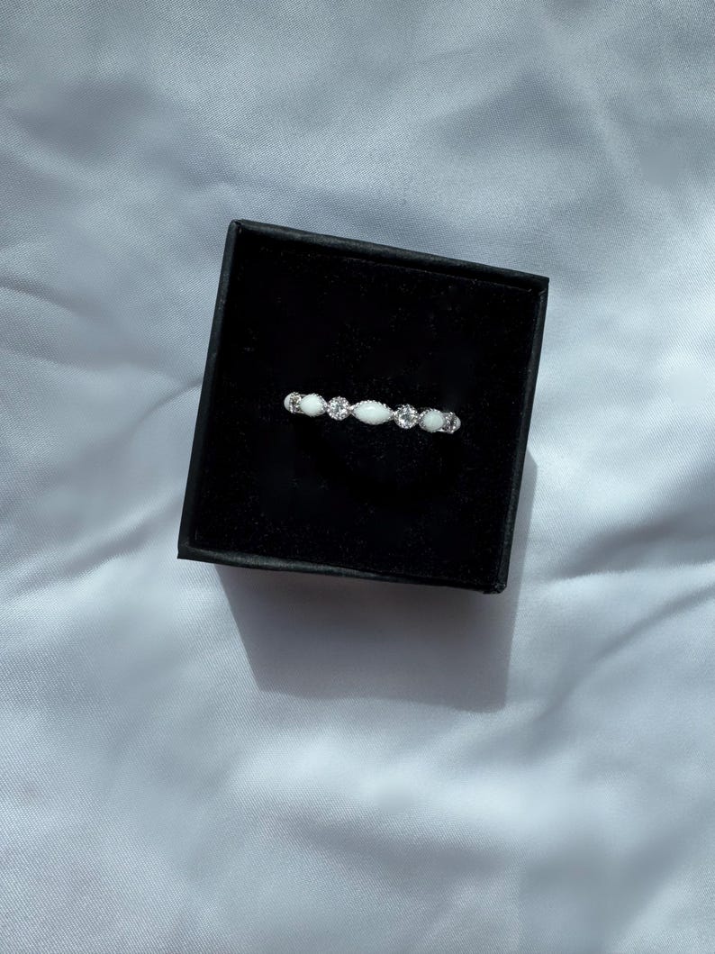 May include: A delicate silver ring with a series of small, clear gemstones and oval-shaped white stones. The ring is displayed inside a black jewelry box, set against a white fabric background. The ring's design is symmetrical and elegant.
