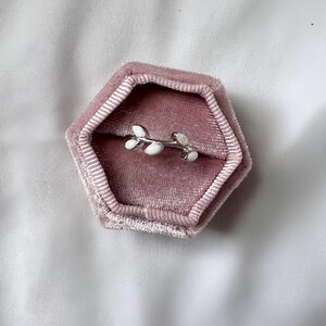 May include: A silver-colored ring with a leaf design, featuring white enamel leaves. The ring is displayed inside a pink velvet hexagonal jewelry box, set against a white satin background. The ring is a delicate piece of jewelry.
