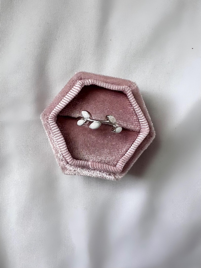 May include: A silver-colored ring with a leaf design, featuring white enamel leaves. The ring is displayed inside a pink velvet hexagonal jewelry box, set against a white satin background. The ring is a delicate piece of jewelry.