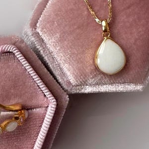 May include: A gold necklace featuring a teardrop-shaped white stone pendant on a delicate chain, positioned alongside a gold ring with small white stones, both resting on a soft pink velvet surface with a hexagonal structure.