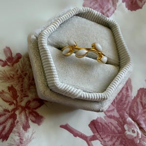 May include: A gold-colored ring with a branch design featuring white, oval-shaped accents. The ring is displayed inside a gray, velvet-lined jewelry box. The box is open, revealing the ring against a floral patterned background.