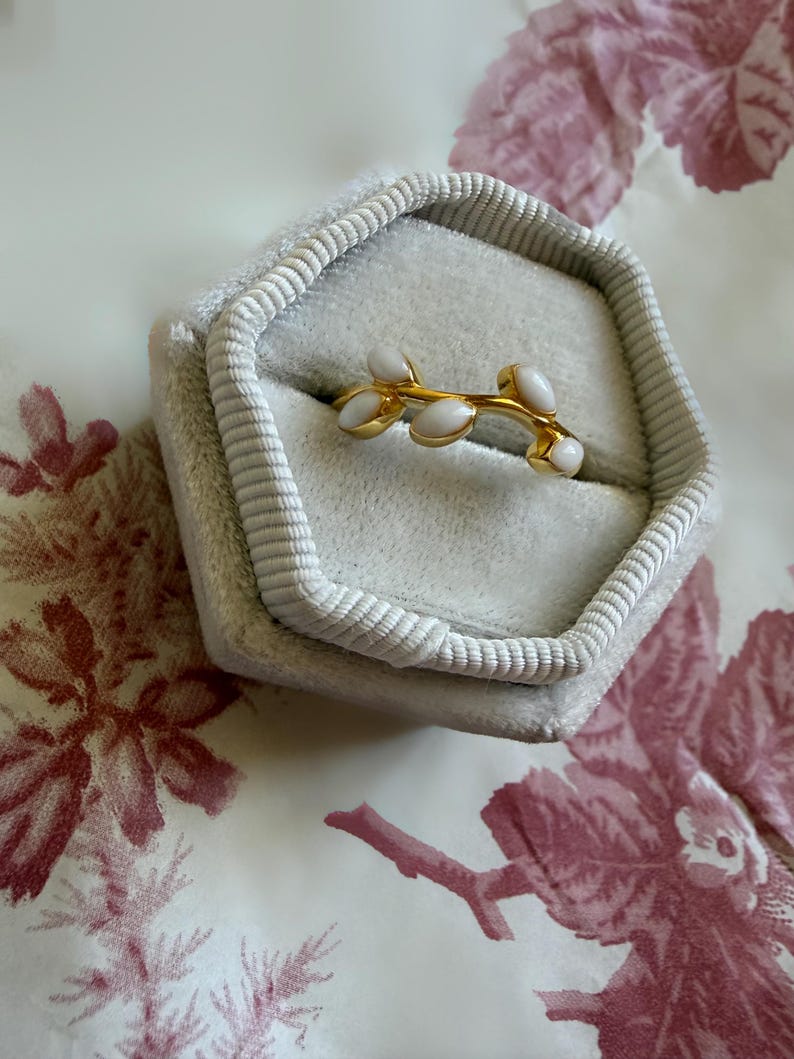 May include: A gold-colored ring with a branch design featuring white, oval-shaped accents. The ring is displayed inside a gray, velvet-lined jewelry box. The box is open, revealing the ring against a floral patterned background.