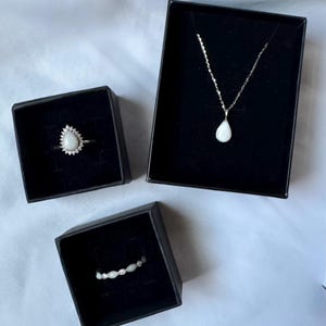 May include: A jewelry set consisting of a teardrop-shaped pendant necklace, a teardrop-shaped ring surrounded by small stones, and a band ring with alternating small white stones, each presented in individual black square gift boxes.