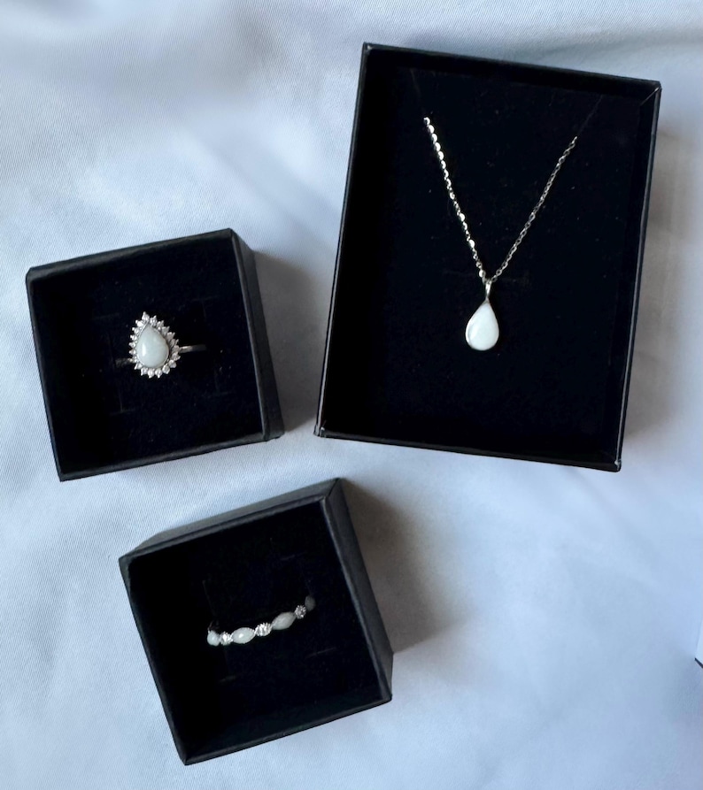 May include: A jewelry set consisting of a teardrop-shaped pendant necklace, a teardrop-shaped ring surrounded by small stones, and a band ring with alternating small white stones, each presented in individual black square gift boxes.