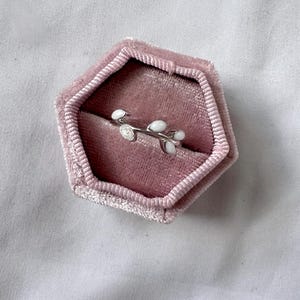May include: A silver ring with a delicate branch design featuring white enamel leaves and a small, clear gemstone. The ring is displayed inside a pink velvet jewelry box, set against a soft, white background.
